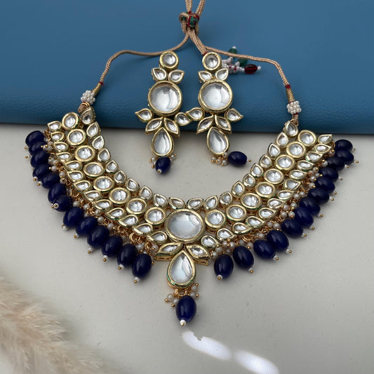 A gold-plated kundan necklace and earring set featuring a floral design. The necklace and earrings have clear, round, and teardrop-shaped accents. Dark blue beads are strung along the necklace's base. The earrings mirror the necklace's design, creating a cohesive jewelry set.