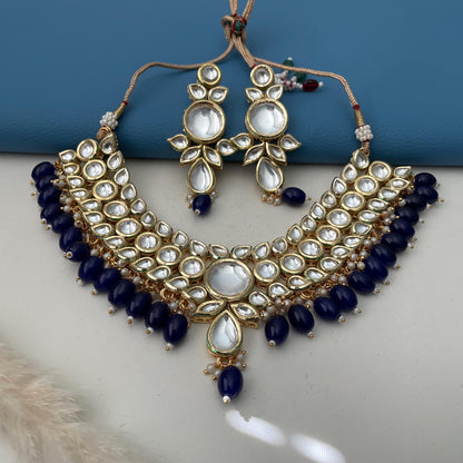 A gold-plated kundan necklace and earring set featuring a floral design. The necklace and earrings have clear, round, and teardrop-shaped accents. Dark blue beads are strung along the necklace's base. The earrings mirror the necklace's design, creating a cohesive jewelry set.