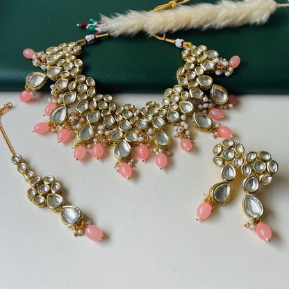 A gold plated  kundan  set featuring a necklace, earrings, and a headpiece. The necklace and earrings have a floral design with clear, teardrop-shaped stones and pink beads. The headpiece mirrors the necklace design. The jewelry is displayed on a white surface.