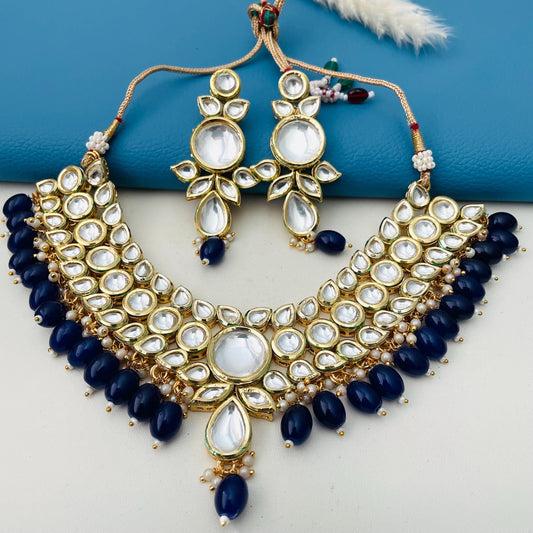 A gold-plated necklace and earring set featuring a bib-style design. The necklace and earrings are adorned with clear, round, and teardrop-shaped kundan, along with navy blue beads. The jewelry set is designed with a traditional Indian aesthetic.