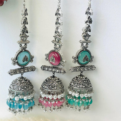 Three silver-colored jhumka earrings with intricate designs. Each earring features a teardrop-shaped pendant with a colored stone in the center, and a bell-shaped bottom adorned with beads. The stones are teal, pink, and mint respectively, with matching bead colors.