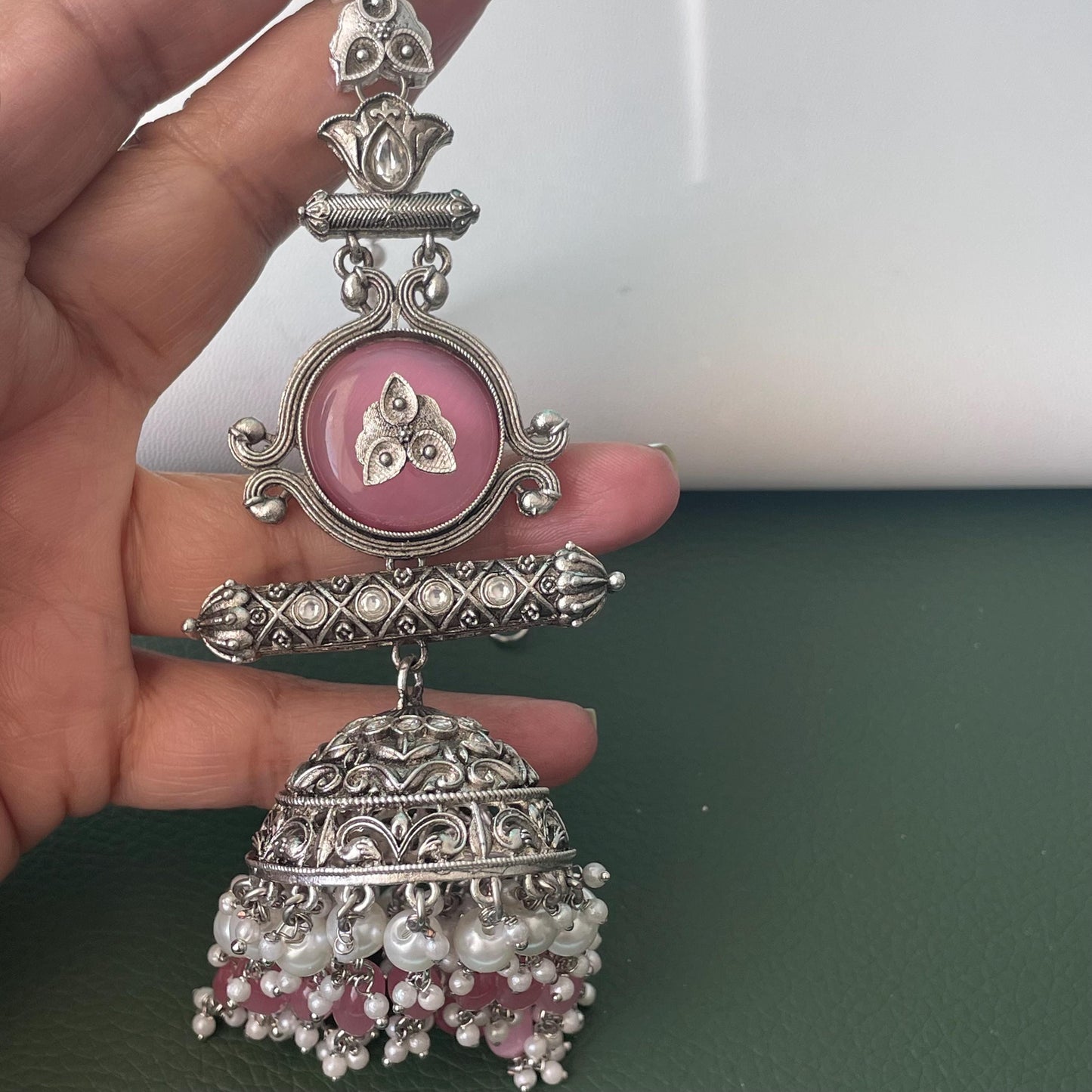 An ornate silver-toned earring with a pink circular center and a dangling bell shape. The earring features intricate detailing, including a floral design on the pink center and a cluster of small white and pink beads at the bottom. The design is symmetrical and traditional, suitable for ethnic or formal wear.