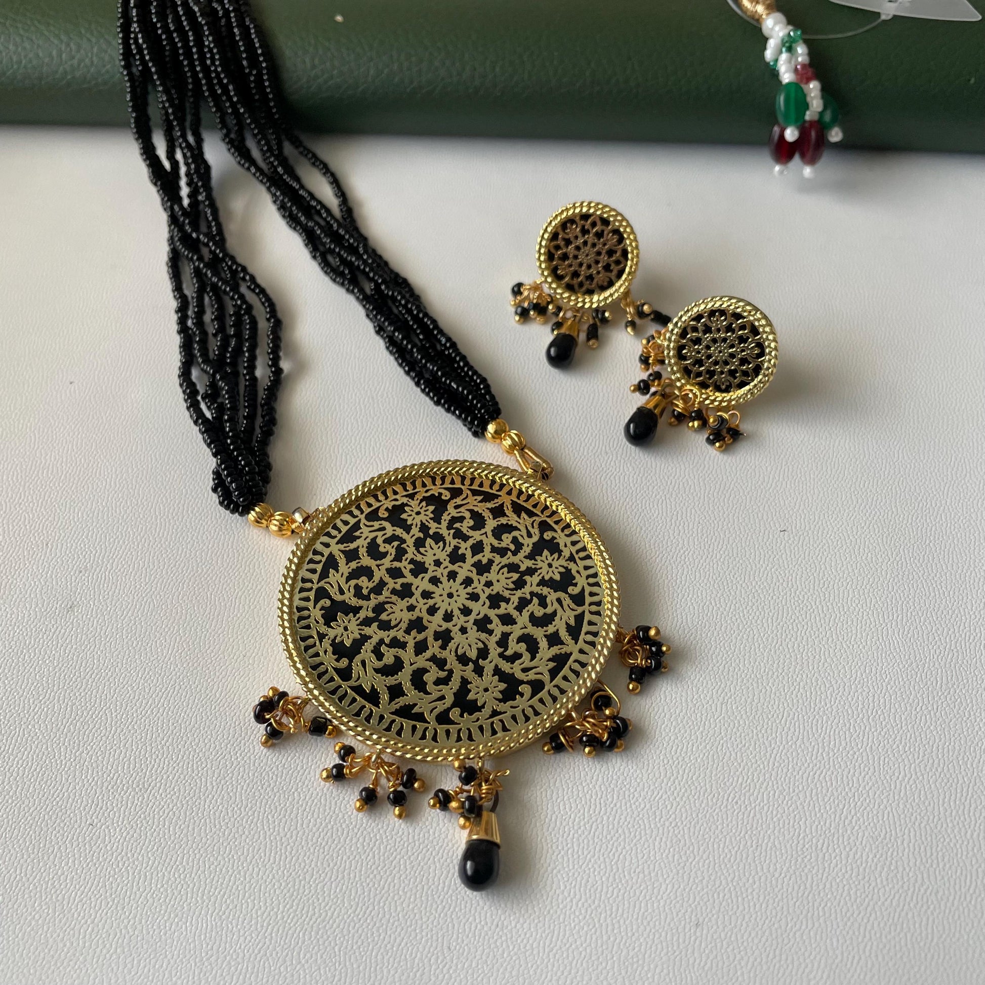 A gold-toned necklace and earring set. The necklace features a large, round pendant with a black background and intricate gold filigree design. The earrings are circular with a similar design. Black beaded strands and accents complete the set. Traditional thewa necklace set