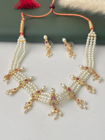 A pearl necklace and earring set. The necklace features multiple strands of white pearls with gold-colored accents and red gemstones. The matching earrings have a similar design. The jewelry is displayed on a white surface. Marathi Chinchapeti
