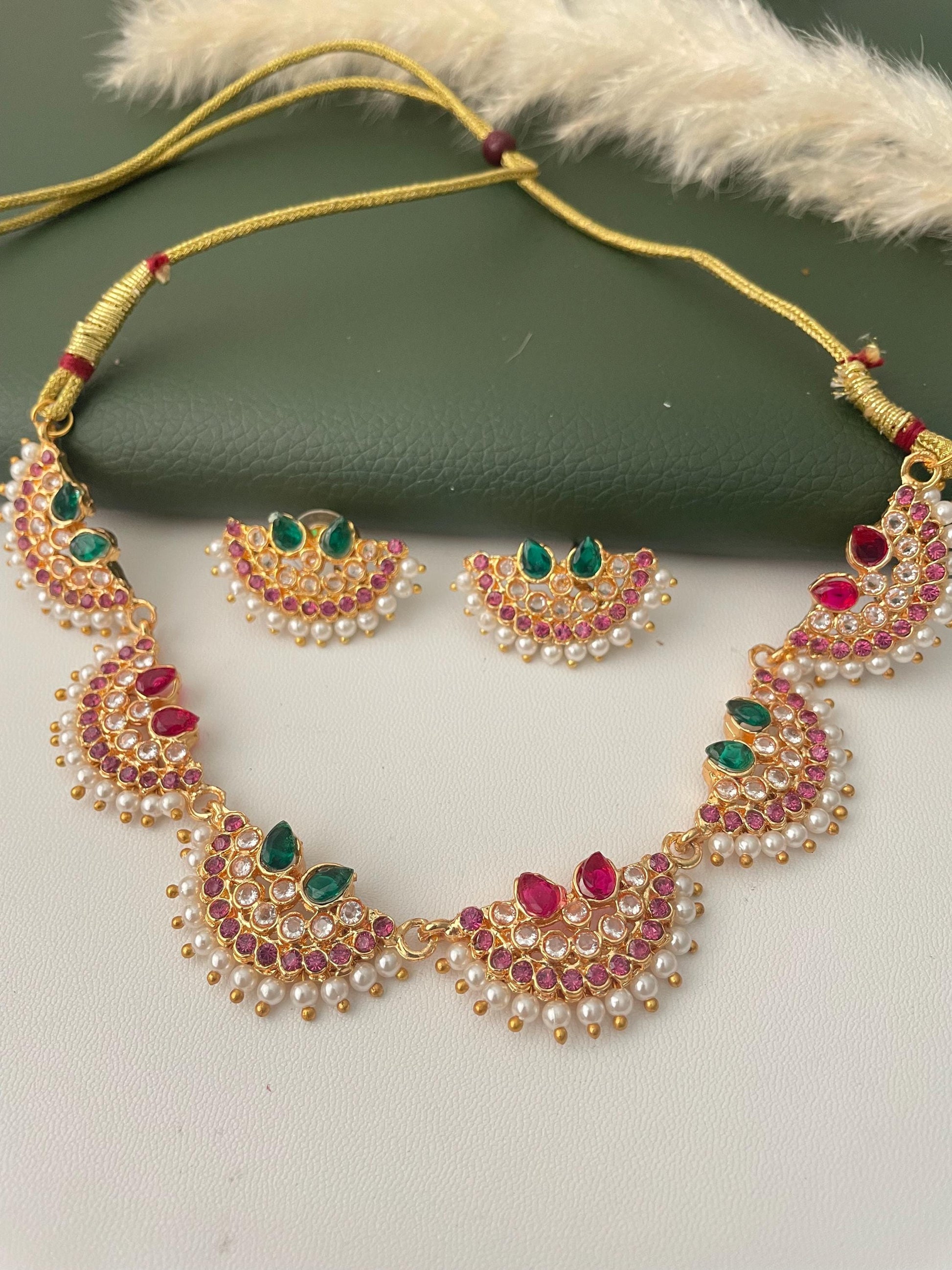 Maharashtrian Chinchapeti: Gold-toned necklace and matching earrings set. The necklace features a series of half-moon pendants adorned with red and green gemstones, surrounded by small white pearls. The earrings mirror the pendant design. The set is displayed on a green surface.