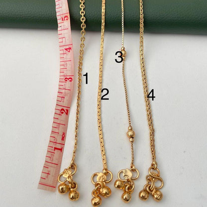 Four gold-colored anklets of varying designs are displayed. The necklaces are measured with a pink and white measuring tape. The necklaces have different chain styles, with some featuring small bell charms.