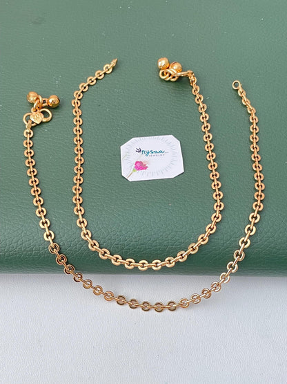 Two gold-colored anklets with a chain link design. Each anklet has a small bell charm and a clasp. The anklets are displayed on a green surface, with a small white tag with text and a flower.