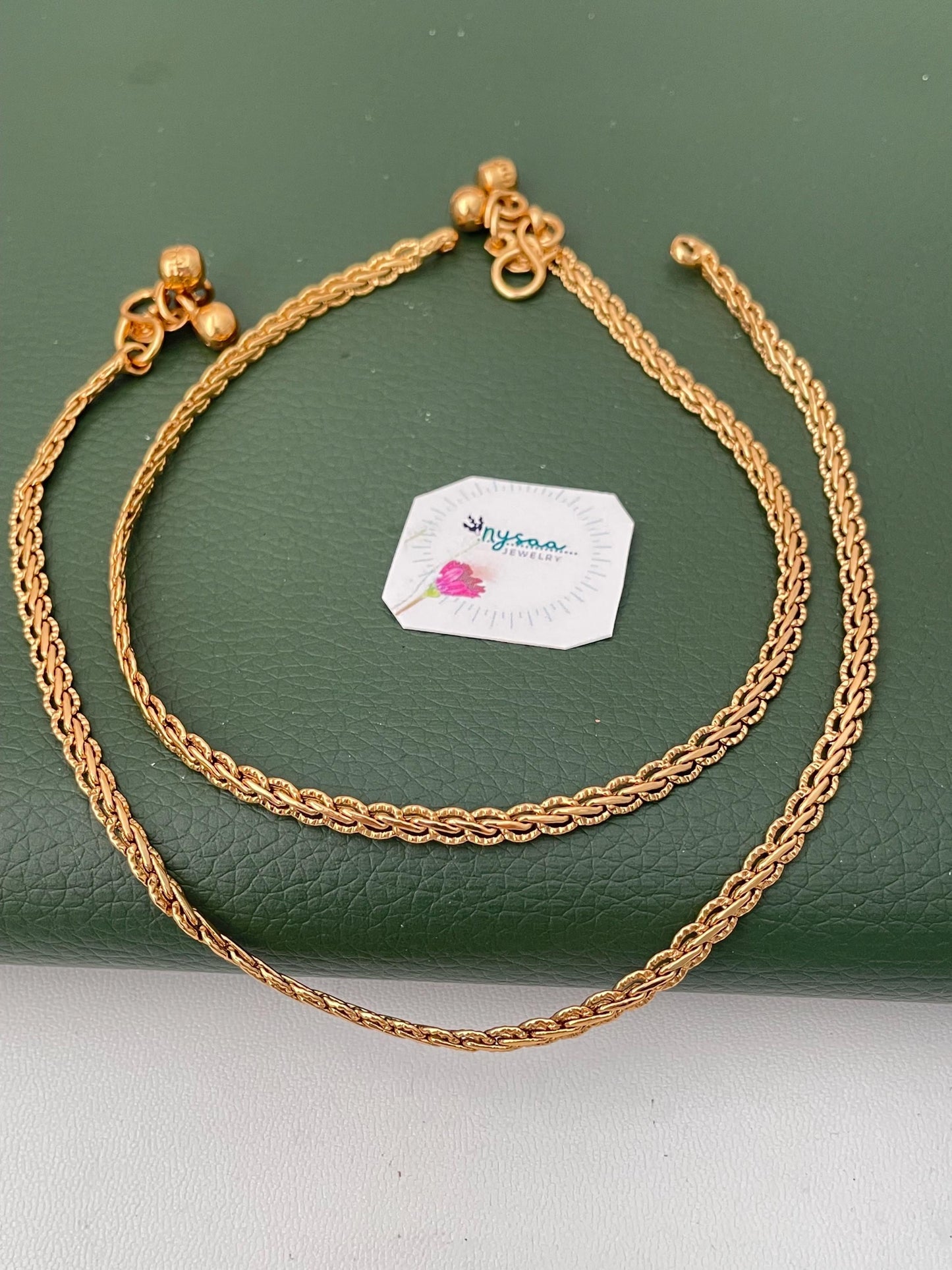 A pair of gold-colored anklets with a delicate chain design. Each anklet features small, dangling bell charms and is displayed on a green surface. The anklets are likely made of a gold-toned metal.