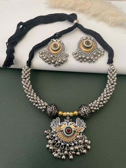 A necklace featuring a black cord, silver-toned beaded segments, and a central pendant. The pendant displays a gold and silver-toned peacock motif with a red and green circular inlay, accented by small dangling silver-toned metal beads.