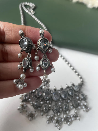 A silver-toned necklace and earring set. The earrings have a teardrop design with clear stones and small white pearls. The necklace features a similar design with multiple clear stones and pearl accents. The jewelry is held against a white and green background.