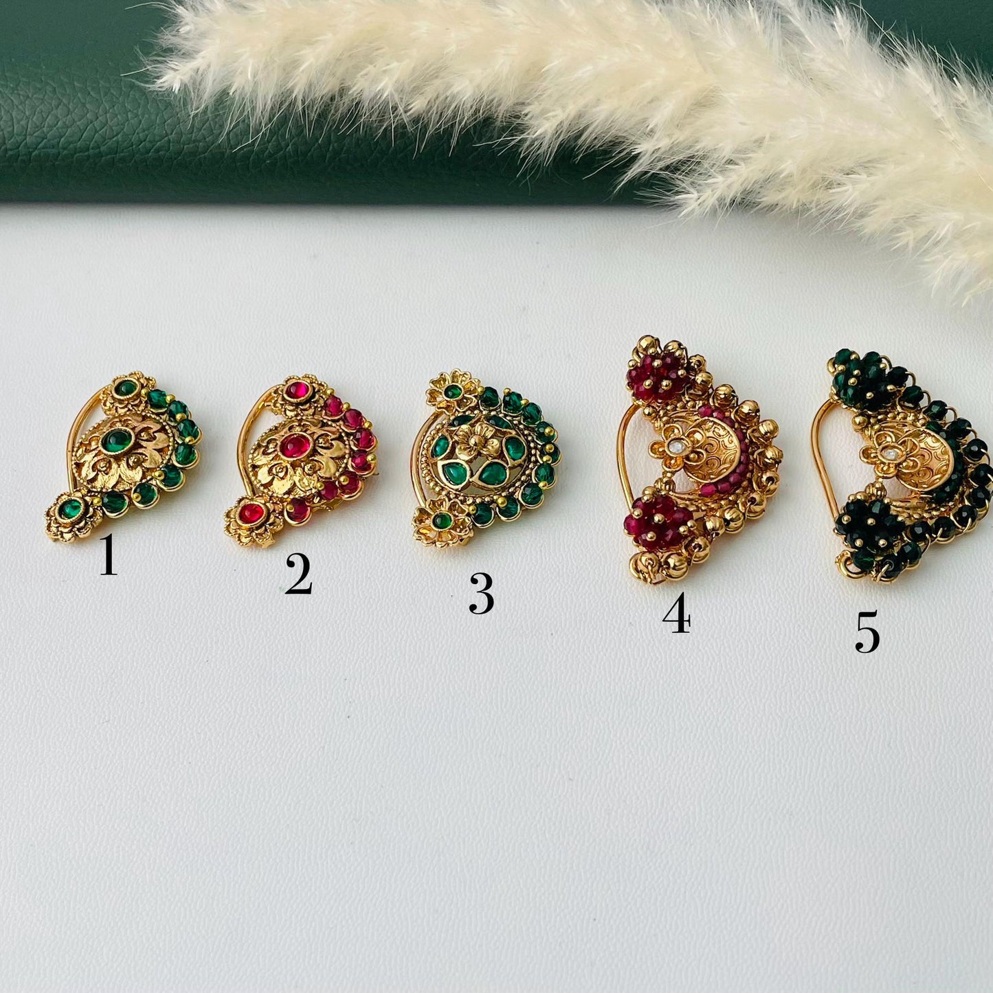 Five ornate gold-toned nose rings with colorful gemstone accents. Designs include green and red stones, with intricate patterns and varying shapes. Each ring is numbered for easy selection.
