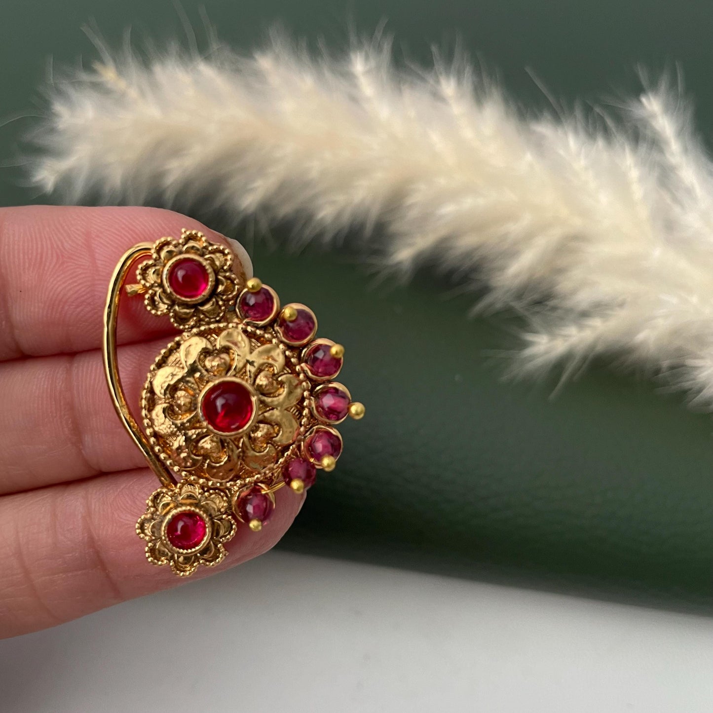 A gold-colored nose ring with a floral design and embedded red gemstones. The ornate jewelry piece features a central flower-shaped element and smaller floral accents, with a curved hook for securing it to the nose.