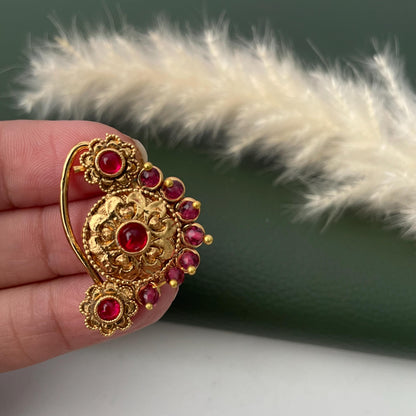 A gold-colored nose ring with a floral design and embedded red gemstones. The ornate jewelry piece features a central flower-shaped element and smaller floral accents, with a curved hook for securing it to the nose.