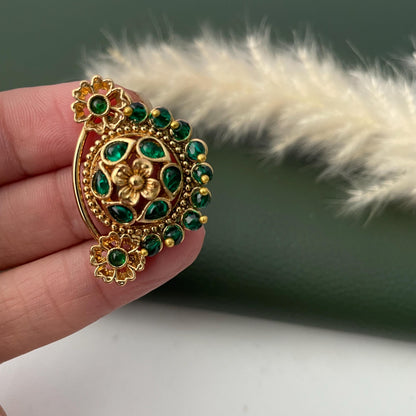 A gold-toned earring with a floral design, featuring a central flower and surrounding green gemstones. The earring has a curved shape with additional floral accents. The design is intricate and detailed, with a focus on the contrast between the gold and green elements.