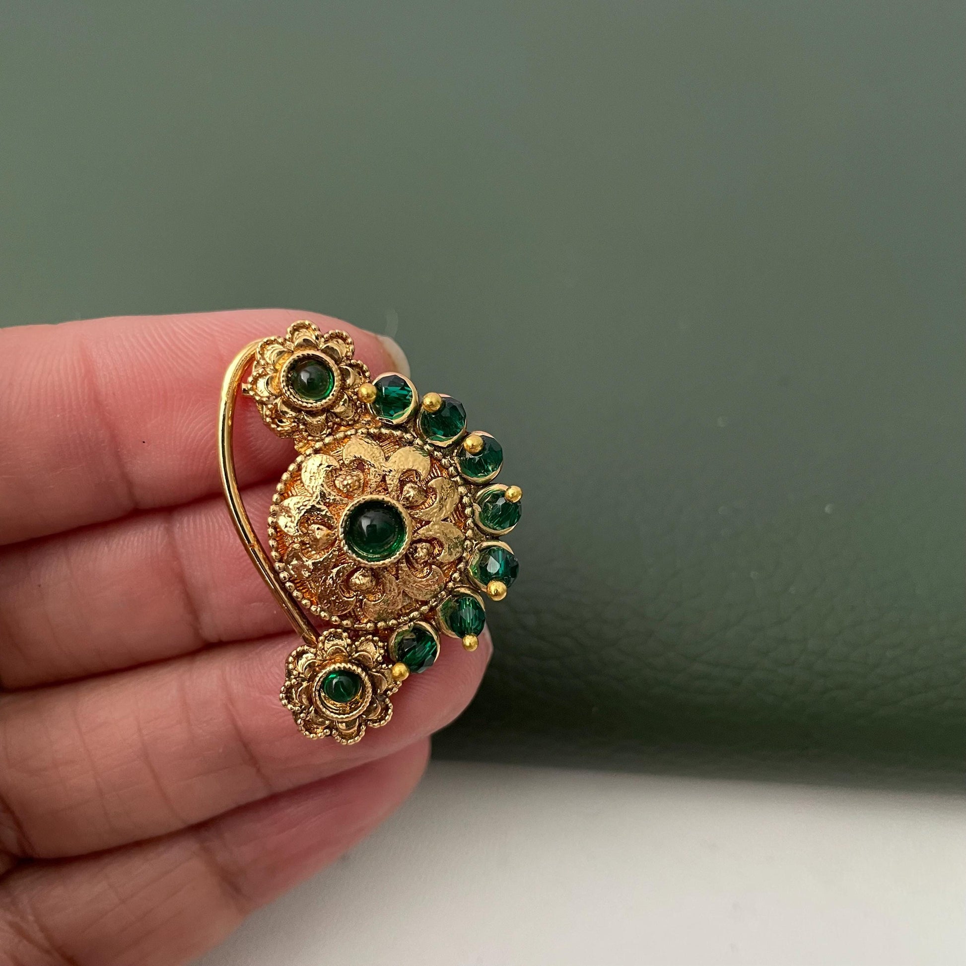 A gold-colored nose ring with a floral design and embedded green gemstones. The jewelry piece has a curved hook for secure placement and features intricate detailing around the central stone and surrounding gems.