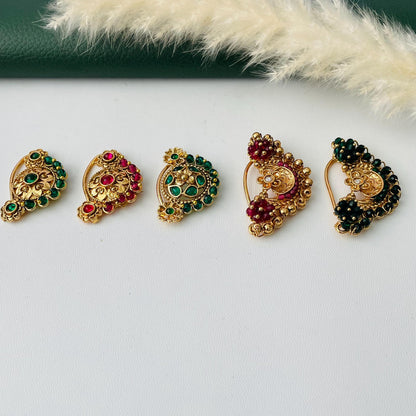 Five ornate gold-colored nose rings with intricate designs. Each ring features a different arrangement of colorful gemstones, including green and red stones. The nose rings are displayed on a white surface, with a green background and a sprig of dried flowers.
