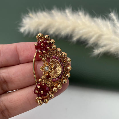 A gold-toned nose ring with a floral design and red accents. The jewelry piece features a curved bar for securing to the nose, with clusters of red beads and a central floral motif with a small, clear stone.