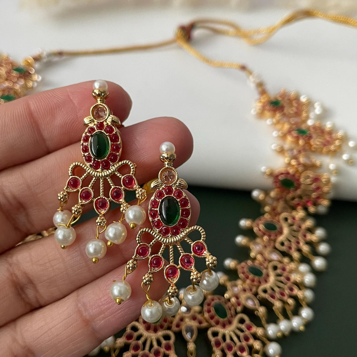 Gold-plated jewelry set featuring earrings and a necklace. The earrings have a chandelier design with red and green gemstones, and dangling pearls. The necklace has a similar design with red and green gemstones and pearls.