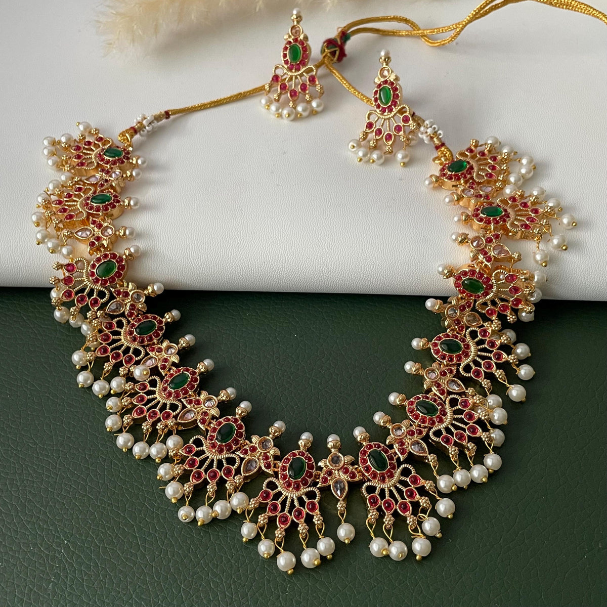 A gold-plated necklace and matching earrings set. The jewelry features red and green gemstones, small white pearls, and intricate detailing. The necklace and earrings are displayed on a white and green surface.