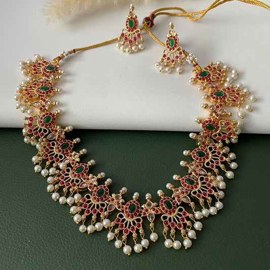 A gold-plated necklace and matching earrings set. The jewelry features red and green gemstones, small white pearls, and intricate detailing. The necklace and earrings are displayed on a white and green surface.