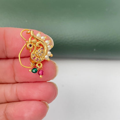 A gold-colored nose ring with a peacock design. The jewelry features small white pearls, red and clear gemstones, and green and pink dangling beads. The ring is held by a person's fingers.