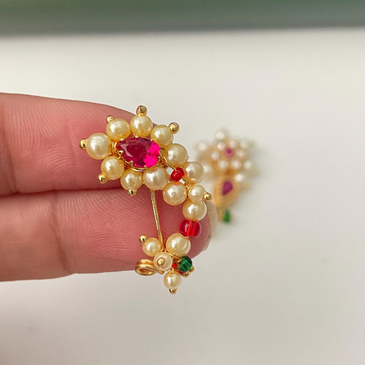 A gold-toned nose ring featuring a central red gemstone surrounded by small, creamy pearls. Additional red and green beads accent the design. The nose ring is designed with a curved shape and a pin for secure placement.