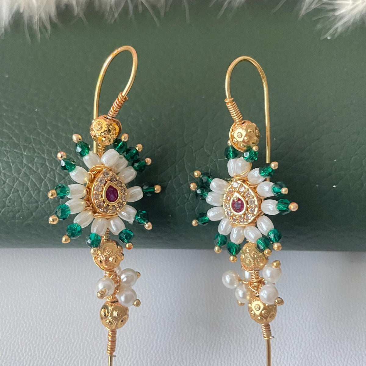 A pair of gold-tone middle earcuffs featuring a central teardrop-shaped charm with a small red stone, surrounded by white pearls and green beads. The earrings include textured gold spheres and long, curved wire hooks for attachment.