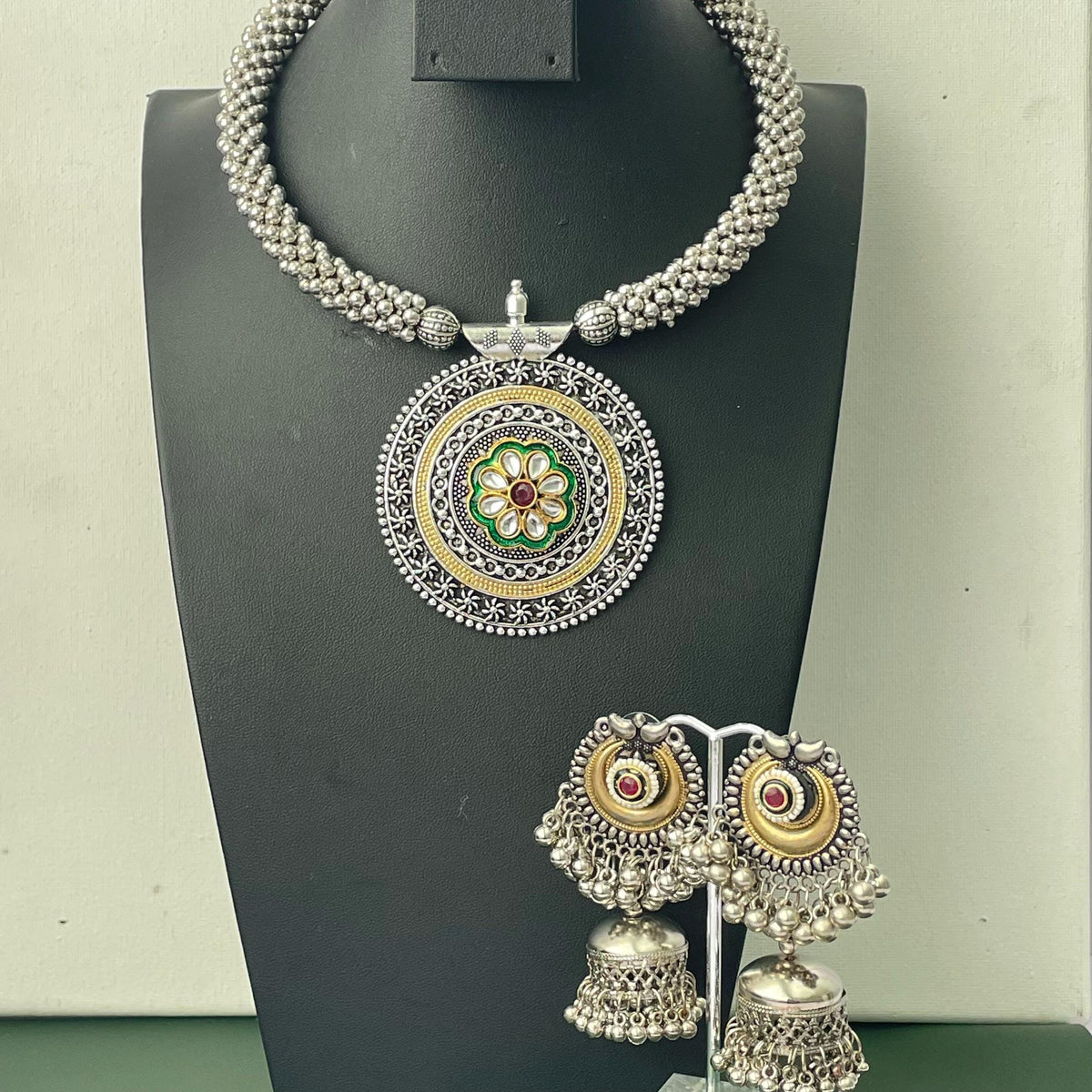 A silver-toned beaded necklace featuring a large circular pendant with gold-tone accents, a central green floral motif, and a small red stone. The set includes two matching jhumka-style earrings with silver beads and red stones.