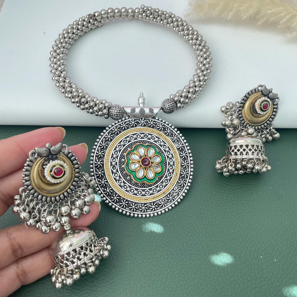 A silver-toned necklace with a beaded chain and a large circular pendant featuring a central green and gold floral motif with a red stone. The set includes two matching jhumka earrings with silver beads and a central red stone.