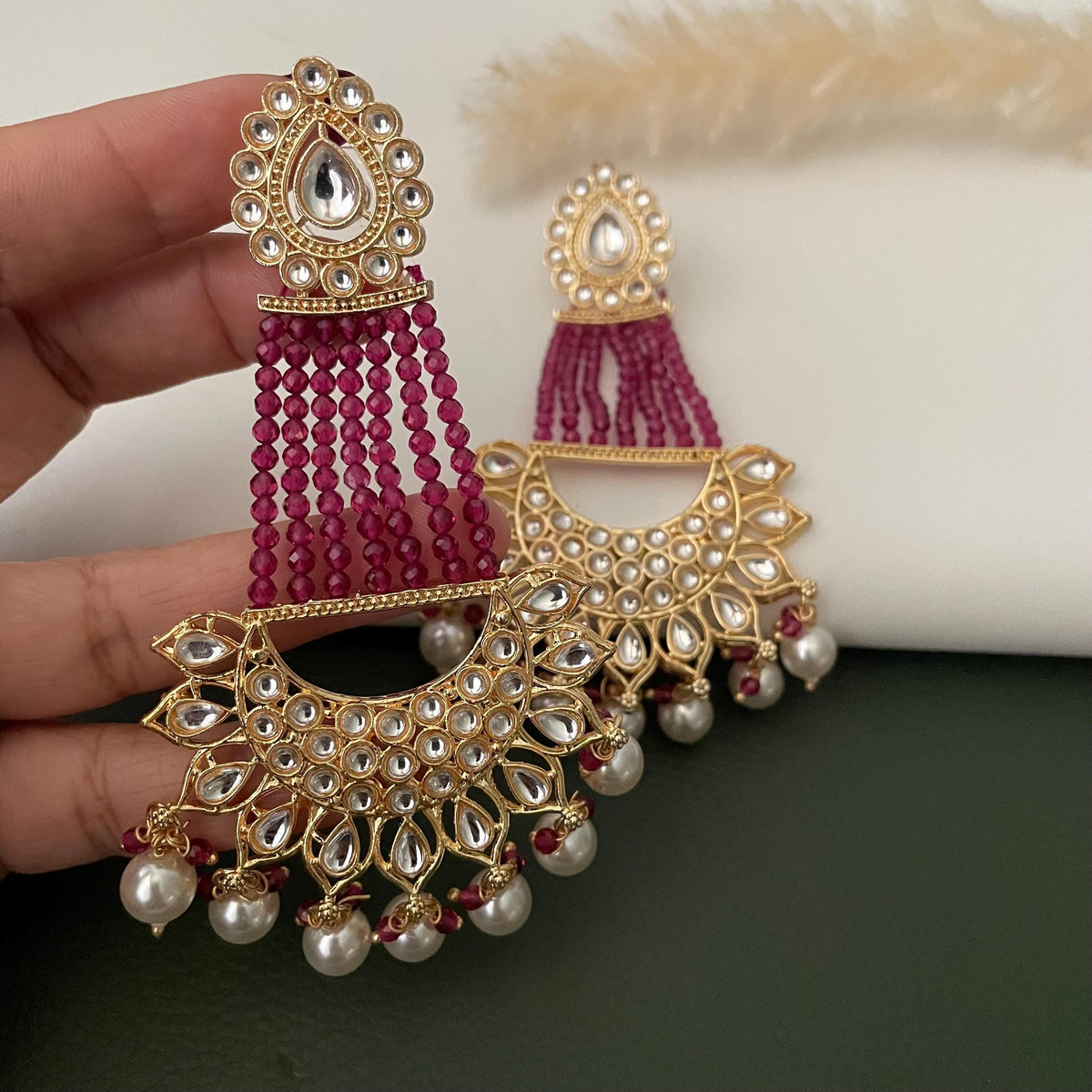 Gold earrings with a crescent moon design, adorned with clear Kundan and green beads. The earrings feature a dangling pearl and maroon bead drop.