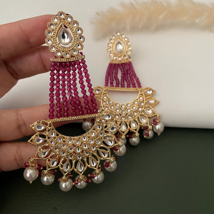 Gold earrings with a crescent moon design, adorned with clear Kundan and green beads. The earrings feature a dangling pearl and maroon bead drop.
