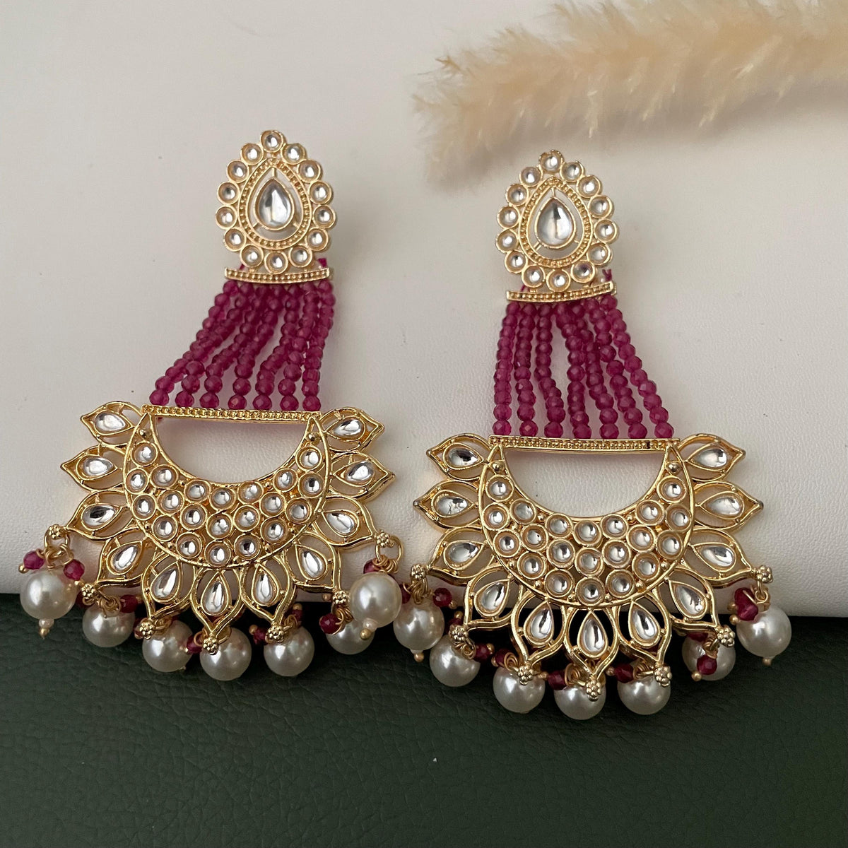 A pair of gold earrings with a teardrop-shaped design, featuring a maroon beaded fringe and white accents.